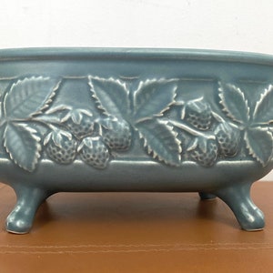 May include: A blue ceramic planter with a raised design of strawberries and leaves. The planter has three legs and is oval shaped.
