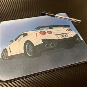 Puede incluir: Alfombrilla de ratón con una ilustración de un coche deportivo blanco. El coche tiene luces traseras rojas y un alerón negro. Un bolígrafo negro descansa sobre la alfombrilla. El fondo es un degradado de azul y gris.