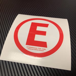 May include: A white sticker featuring a red circular border and a bold red letter "E". The text "stormvaultuk.com Motorsport Graphics Specialist" is printed at the bottom of the sticker.