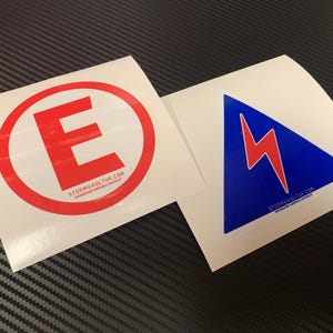 Könnte beinhalten: Zwei weiße Aufkleber. Einer zeigt ein rotes "E" in einem roten Kreis, der andere einen roten Blitz in einem blauen Dreieck. Beide haben die Website stormvaultuk.com unten aufgedruckt.