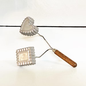 May include: Vintage metal cookie cutters in heart and square shapes, attached to a wire frame with a wooden handle. The cutters have a scalloped edge and are made of a silver-colored metal. The handle is a dark brown color.