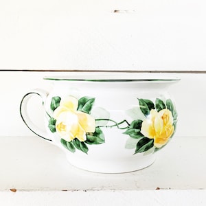 May include: White ceramic chamber pot with a green rim and handle, decorated with yellow roses and green leaves. The floral design is painted on the exterior of the pot, creating a vintage aesthetic.