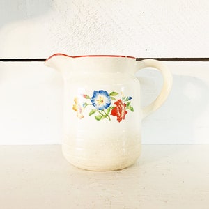 May include: Cream-coloured ceramic jug with a red rim and a handle. The jug is decorated with a floral design featuring blue, red, and yellow flowers with green leaves. The jug is on a white surface.