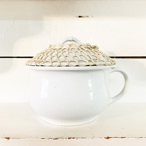 Puede incluir: Olla de cerámica blanca con asa y tapa de ganchillo. La tapa beige tiene un asa circular. La olla está sobre una estantería blanca. El estilo es rústico y vintage.