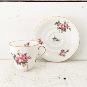 Puede incluir: Un juego de taza y platillo blancos con un estampado de rosas rosas y verdes. La taza y el platillo tienen ribetes dorados. El juego se exhibe sobre una superficie de madera blanca.