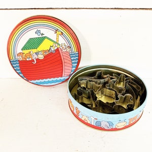 Könnte beinhalten: Ein Vintage-Ausstechersatz in einer runden Dose. Der Deckel zeigt eine farbenfrohe Illustration der Arche Noah mit Tieren und einem Regenbogen. Die Dose enthält verschiedene Metallausstecher in verschiedenen Formen.