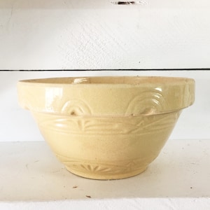 Puede incluir: Un cuenco mezclador vintage de color amarillo claro con un borde ancho y patrones decorativos en relieve. El cuenco tiene una forma clásica y está hecho de cerámica. El diseño incluye patrones curvos y lineales alrededor del borde y la base.