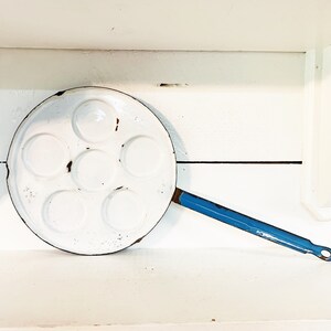 Puede incluir: Una sartén para freír huevos vintage de esmalte blanco con siete moldes circulares. La sartén tiene un mango azul y muestra signos de desgaste, lo que sugiere su antigüedad. Es un artículo de menaje.
