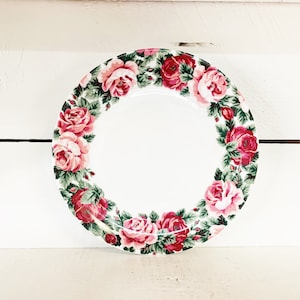 Puede incluir: Un plato de cerámica blanco con un diseño floral rosa y verde. El plato tiene un centro blanco y un borde de rosas rosas y hojas verdes.