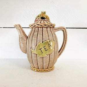 Puede incluir: Una tetera de cerámica con un diseño de cesta tejida y una hoja verde con la palabra "TEA" en ella. La tetera tiene un diseño floral en la tapa.