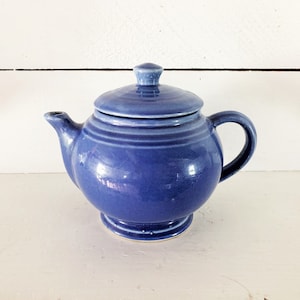May include: A blue ceramic teapot with a matching lid. The teapot has a rounded body, a curved handle, and a spout. The lid has a small knob on top. The teapot is sitting on a white surface.
