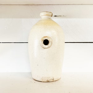 Puede incluir: Una jarra de cerámica blanca con un agujero redondo en el lateral. La jarra tiene un esmalte marrón y el texto "DORCHESTER POTTERY BOSTON MASS" en la parte inferior.