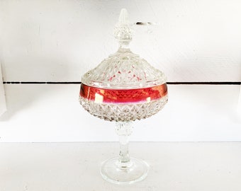 Puede incluir: Un bombonera de cristal transparente con un patrón tallado en diamante y una banda roja iridiscente. La tapa está rematada con un adorno decorativo. El recipiente está sobre un pie y sobre una superficie blanca.