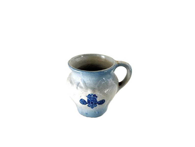May include: A small, light blue and white ceramic pitcher with a handle. The pitcher has a decorative blue floral design on the front. The interior of the pitcher is a darker gray color.