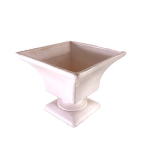 Könnte beinhalten: Ein weißer Keramik-Pflanztopf mit quadratischer Oberseite und Sockel. Der Pflanztopf hat ein ausgestelltes, leicht gebogenes Design. Die Gesamtform ist geometrisch und symmetrisch, geeignet für die Präsentation von Pflanzen oder Blumen.