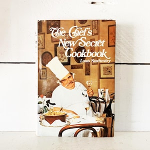 May include: Vintage cookbook titled "The Chef's New Secret Cookbook" by Louis Szathmary. The cover features a chef in a white uniform and hat, holding a glass of wine. The background shows framed pictures and a dining scene.