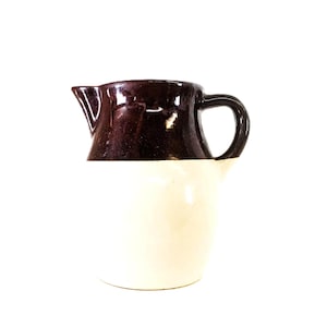 May include: A small, two-toned ceramic pitcher. The top half is a dark brown, and the bottom half is cream-colored. It has a handle and a spout for pouring. The pitcher is likely for serving liquids.