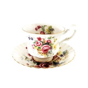 May include: A white porcelain teacup and saucer set featuring a delicate floral pattern with pink roses in a woven basket, green leaves, and small dark berries. The rim of both the cup and the saucer is finished with a thin gold metallic trim.