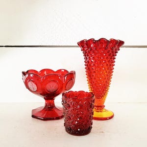 Op de afbeelding: Drie stukken rode glazen woondecoratie. Een voetkom met een geschulpte rand, een kleine tumbler en een hoge vaas met een gerimpelde bovenkant. De vaas heeft een textuur en een gele basis. De objecten zijn op een wit oppervlak geplaatst.