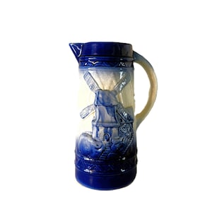 Puede incluir: Una jarra de cerámica con un diseño en azul y blanco que presenta un relieve de un molino de viento en la parte frontal. El recipiente tiene un borde y una base de color azul oscuro, un asa curva y un pico. El cuerpo central muestra un molino.