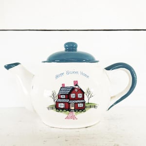 Puede incluir: Tetera de cerámica blanca con tapa y asa azules. La tetera presenta una ilustración de una casa roja con las palabras "Home Sweet Home" en escritura azul. La tetera está diseñada para preparar té.