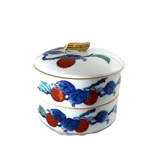 Puede incluir: Un tarro de cerámica blanco con tapa y diseño de dos niveles. El tarro está decorado con fruta roja y hojas azules. La tapa tiene un asa dorada. El tarro se utiliza probablemente para guardar alimentos u otros objetos pequeños.
