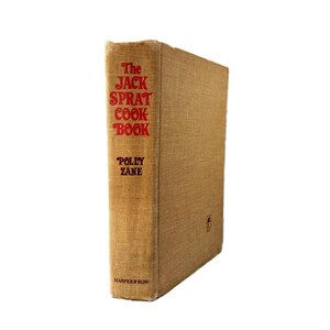 May include: A vintage cookbook titled "The Jack Sprat Cook Book" by Polly Zane. The book has a tan linen cover with red lettering and a small emblem. The publisher's name, Harper & Row, is printed at the bottom.