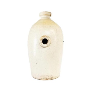 May include: A cream-colored stoneware jug with a cylindrical body, a narrow neck, and a circular opening on the front side. The base features stamped text that reads "BOSTON MASS". The surface has a speckled texture and a smooth, glazed finish.