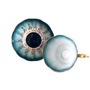 May include: A teal and white porcelain teacup and saucer set featuring a scalloped edge design. The saucer displays intricate gold filigree patterns around a dark teal center, while the teacup has a white interior and a gold-trimmed handle.