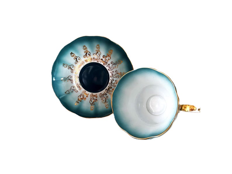May include: A teal and white porcelain teacup and saucer set featuring a scalloped edge design. The saucer displays intricate gold filigree patterns around a dark teal center, while the teacup has a white interior and a gold-trimmed handle.