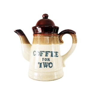 Puede incluir: Una cafetera de cerámica con base color crema y tapa, pico y asa de color marrón oscuro. El cuerpo presenta una textura moteada y el texto azul oscuro COFFEE FOR TWO impreso en la parte frontal. La cafetera tiene una base ancha y asa curva.