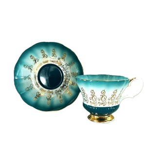 May include: A teal and white porcelain teacup and saucer set featuring a scalloped edge design. The pieces are decorated with intricate gold filigree patterns and a solid teal base, finished with a gold-trimmed handle and a gold-painted footed base.