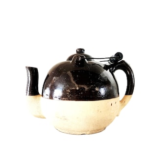 Puede incluir: Una tetera de cerámica redonda con un diseño de dos tonos, que presenta un esmalte marrón oscuro brillante en la mitad superior y una base mate de color crema. Incluye un pico curvo, una tapa a juego con un pequeño pomo y un asa oscura.