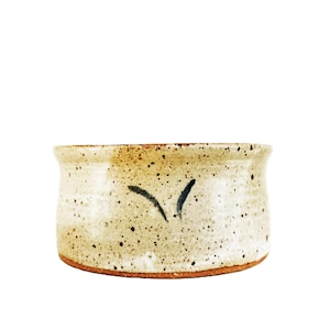 May include: A handcrafted ceramic bowl with a speckled, off-white glaze. The bowl has a wide, shallow shape with a dark brown rim and base. A simple, dark blue leaf design is painted on the side.