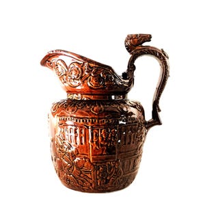 May include: A brown ceramic pitcher with intricate relief details. The pitcher features a detailed handle with a horse head design, a spout with floral patterns, and scenes of people and buildings around the body.
