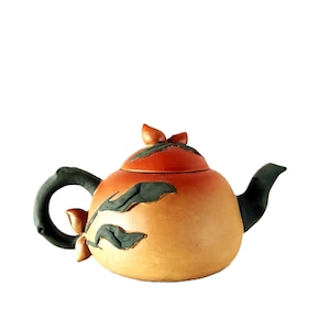Może przedstawiać: Ceramiczny czajnik w odcieniach pomarańczu i beżu, z ciemnozielonymi motywami liści i pasującym uchwytem i dzióbkiem. Pokrywka jest ciemnopomarańczowa, a czajnik ma zaokrąglony korpus. Dekoracyjny design.