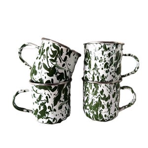 May include: A set of four white enamel mugs featuring a dark green marbled splatter pattern. Each mug has a cylindrical body, a curved handle, and a silver-toned metal rim. The mugs are arranged in a stack of two on the left and two on the right.