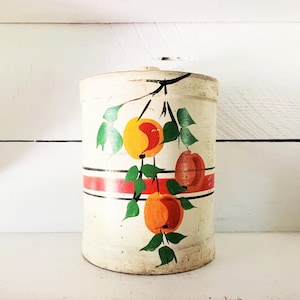 Può includere: Un barattolo cilindrico vintage con base color crema. Presenta frutta arancione e marrone dipinta a mano con foglie verdi. Una striscia rossa è centrata sul barattolo, con strisce nere sopra e sotto.