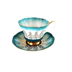 May include: A porcelain teacup and saucer set featuring a scalloped edge, a gradient teal to white finish, and intricate gold filigree scrollwork patterns. The cup has a gold-trimmed handle and a gold-painted base, matching the saucer's design.