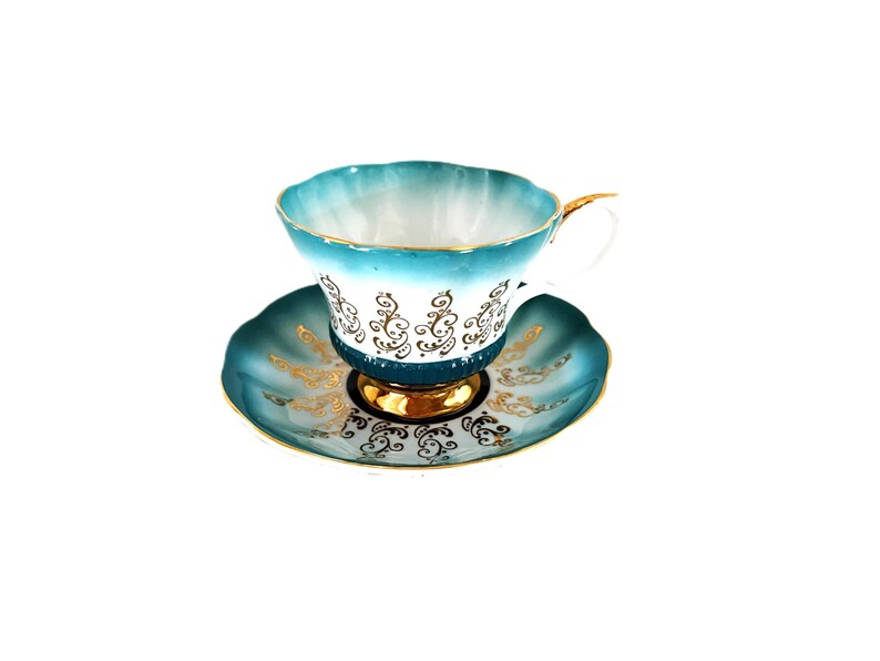 May include: A porcelain teacup and saucer set featuring a scalloped edge, a gradient teal to white finish, and intricate gold filigree scrollwork patterns. The cup has a gold-trimmed handle and a gold-painted base, matching the saucer's design.