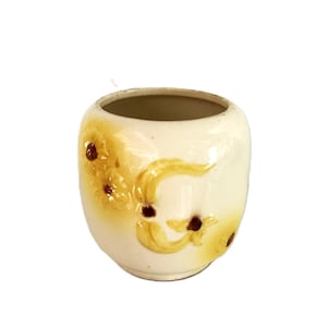 May include: A cream-colored ceramic planter with a rounded, bulbous shape. The exterior features embossed, raised yellow floral designs with dark brown centers. The surface has a glossy finish, and the interior is a smooth, light beige color.