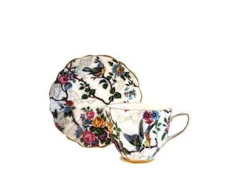 Puede incluir: Un juego de taza de té y plato de porcelana blanca con un patrón floral colorido de flores rosas, amarillas y azules, hojas verdes y pequeños pájaros posados en ramas. Los bordes del plato y el asa de la taza tienen un acabado dorado.