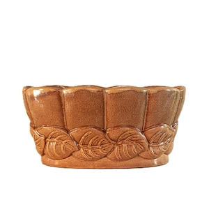 Peut inclure: Un cache-pot en céramique beige avec une finition brillante, présentant un bord supérieur festonné et une bande décorative de feuilles en relief autour de la base. Le bol a une forme ovale arrondie et une couleur brun clair uniforme.
