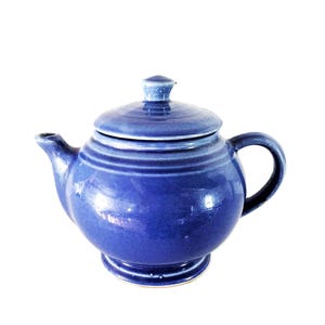 May include: A round, cobalt blue ceramic teapot with a glossy finish. The teapot features a matching lid with a small knob, a curved spout, and a sturdy handle. The body has a wide, bulbous shape that tapers down to a circular pedestal base.