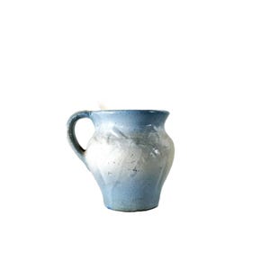 May include: A small, handmade ceramic pitcher with a rounded body and a curved handle. The pitcher is primarily light blue, with a lighter, almost white, area in the middle. The rim is a darker blue.