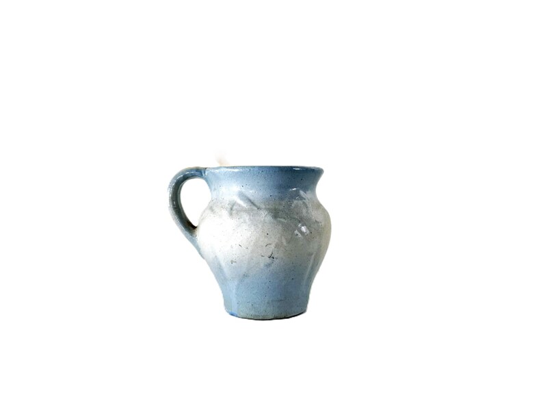 May include: A small, handmade ceramic pitcher with a rounded body and a curved handle. The pitcher is primarily light blue, with a lighter, almost white, area in the middle. The rim is a darker blue.
