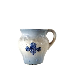 Op de afbeelding: Een vintage keramische kan in lichtblauwe en crèmekleurige tinten. Het heeft een donkerblauw bloemmotief op de voorkant en een gebogen handvat. Deze kan is waarschijnlijk bedoeld voor het serveren van drankjes.