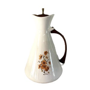 May include: A white conical pitcher featuring a brown floral pattern on the front. The pitcher has a dark brown handle, a matching dark brown lid, and a small gold-toned metal finial at the top of the lid. The body is smooth and glossy.