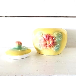 Pode incluir: Um pote de cerâmica amarelo com tampa, com um desenho floral em relevo em rosa e verde. O pote é redondo com uma tampa a condizer, perfeito para guardar pequenos objetos. A tampa tem uma pequena pega. O pote tem cerca de 10 cm de altura.