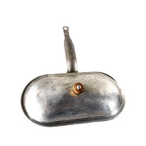 Op de afbeelding: Een ovaalvormig metalen object met een handvat en een kleine, cirkelvormige bevestiging. Het object heeft een zilveren afwerking en lijkt van metaal te zijn. Het handvat heeft een lus aan het uiteinde. Waarschijnlijk een keukengerei.